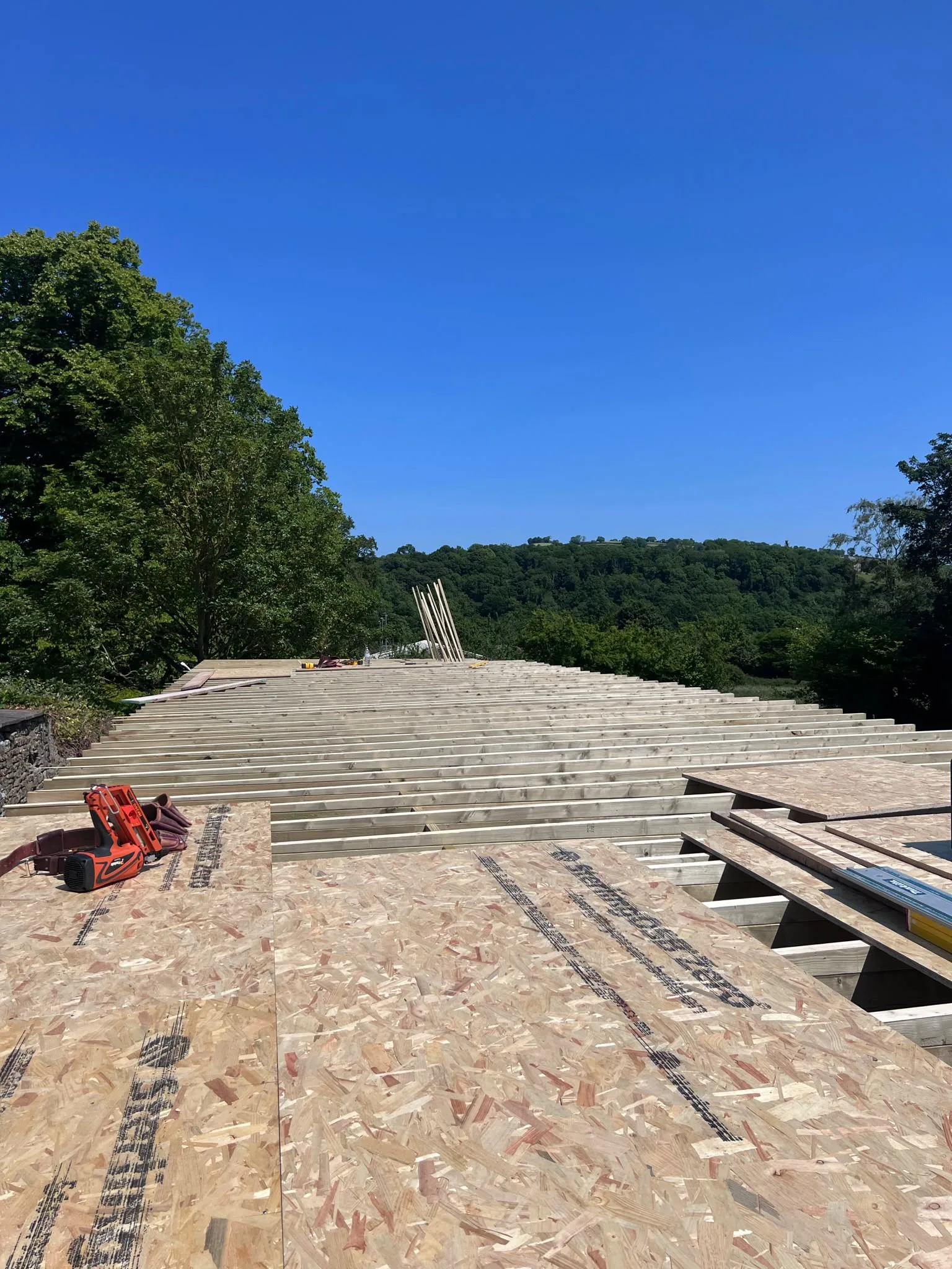 Low-slope roof mid-construction: 4x8 OSB sheathing panels nailed in staggered courses with framing nailer staged on the deck, parallel joists extending to the perimeter beam