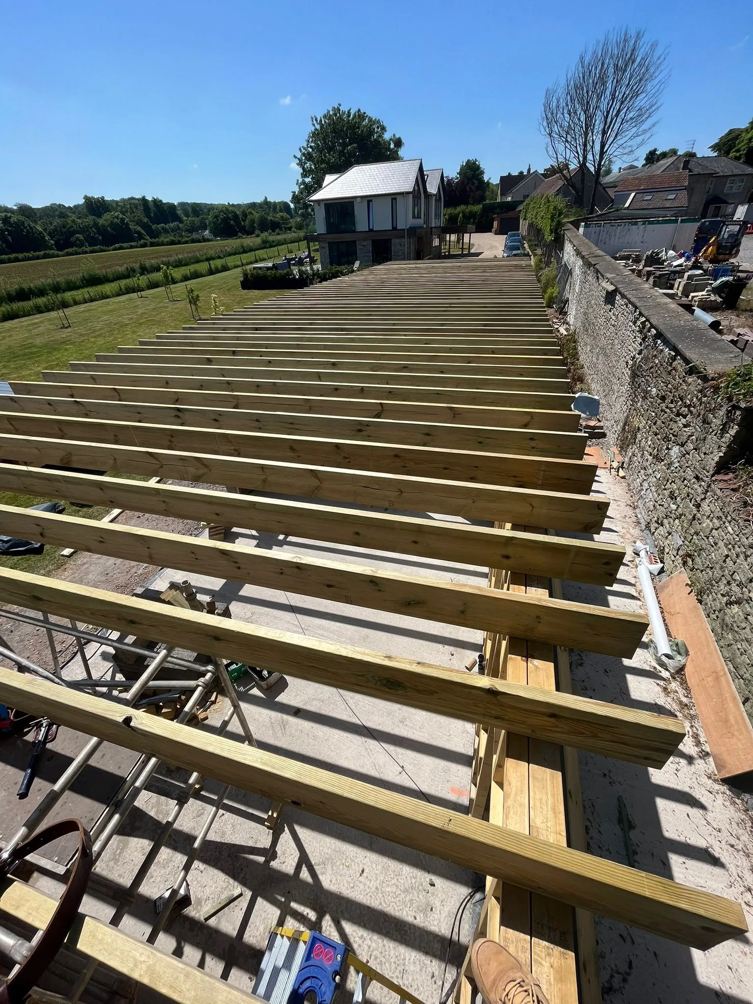 Parallel floor joists on a new-build project running into a retained stone boundary wall, viewed from scaffolding with a modern house in the background
