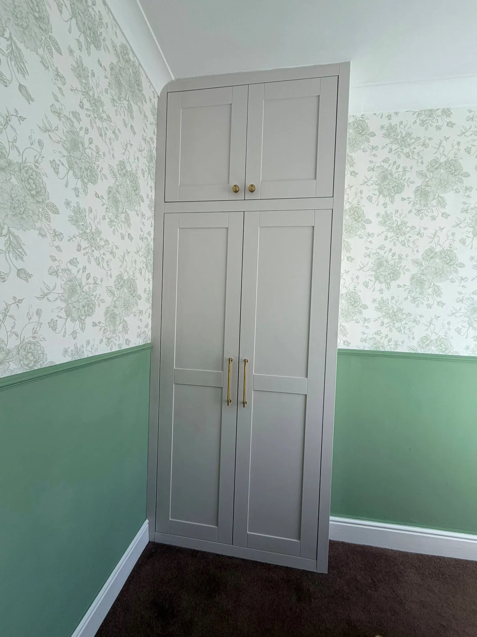 Floor-to-ceiling light grey shaker fitted wardrobe with brass knobs and bar handles, set in a bedroom with sage green lower walls and floral wallpaper
