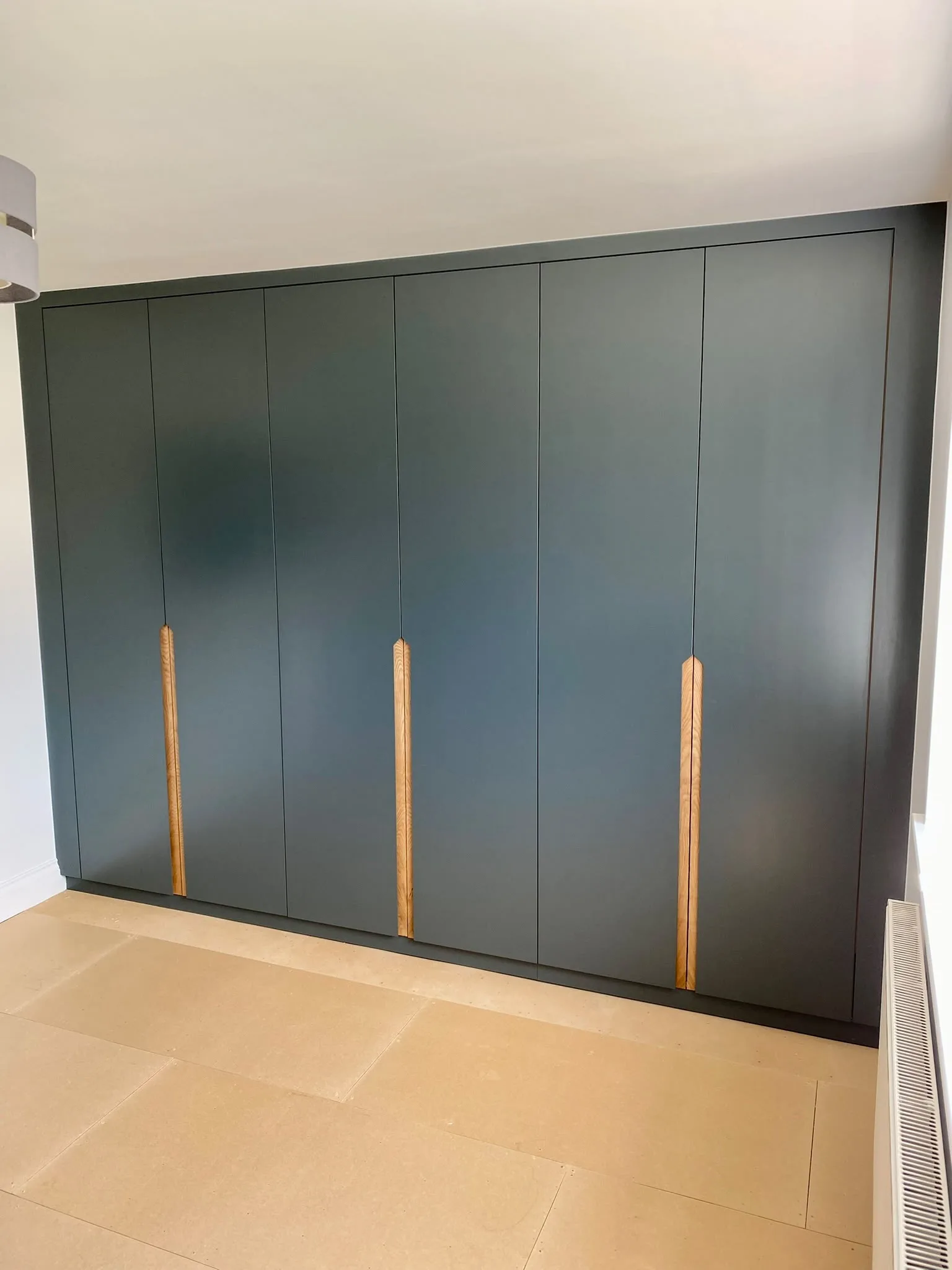 Floor-to-ceiling dark green handleless fitted wardrobe with five flat slab doors and vertical natural oak pull handles, in a minimal cream room with beige tiled floor